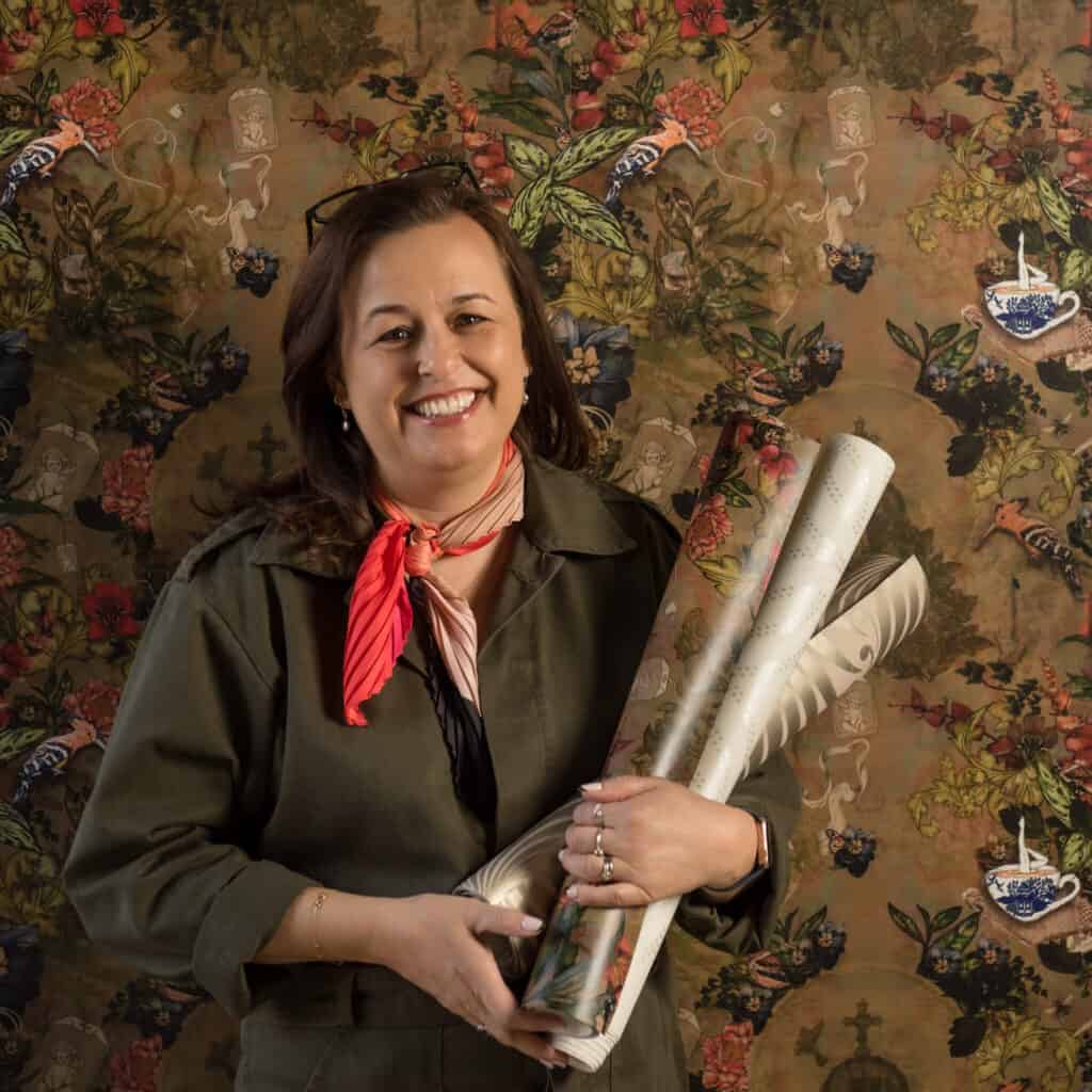 Interior Designer Joanne Jeyes standing in front of a wall featuring a patterned wallpaper. She is holding rolls of wallpaper and smiling