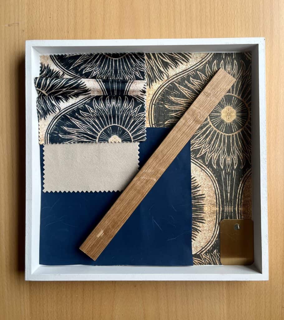 A sample board showing blue and gold fabric, wallpaper, walnut wood, beige and navy velvet fabric all sat within a white tray.